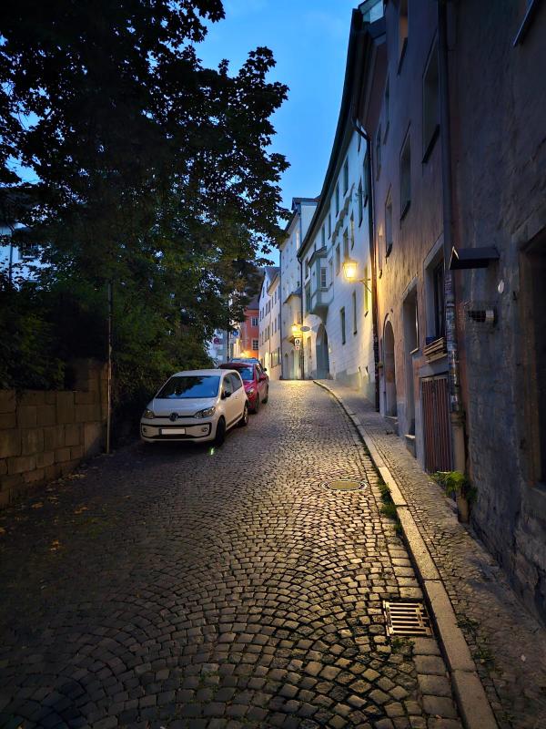 Gasse mit Altstadthäuser rechts, links eine alte Mauer mit Bäumen und parkenden Autos.