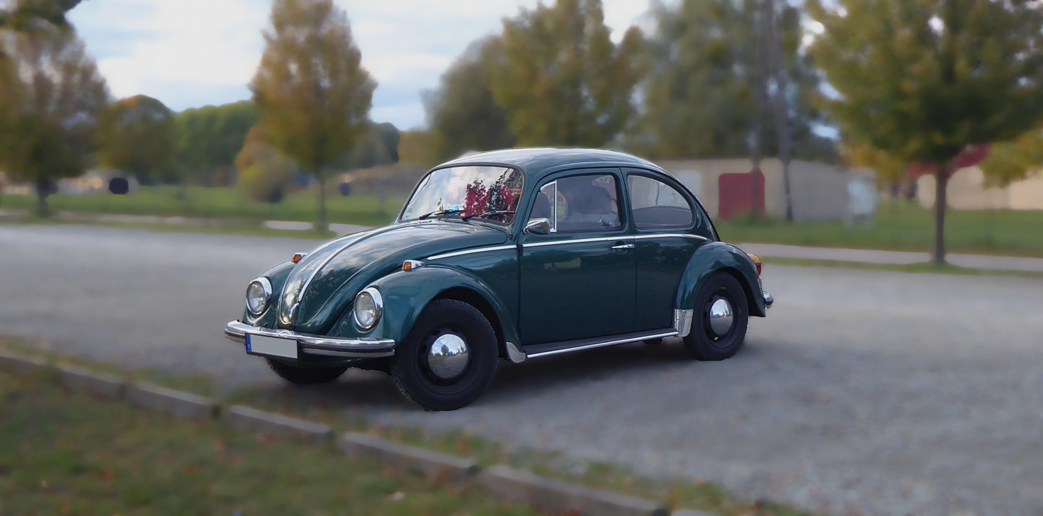 VW-Käfer in grünmetallic auf einem Parkplatz. Der Bildhintergrund ist unscharf.